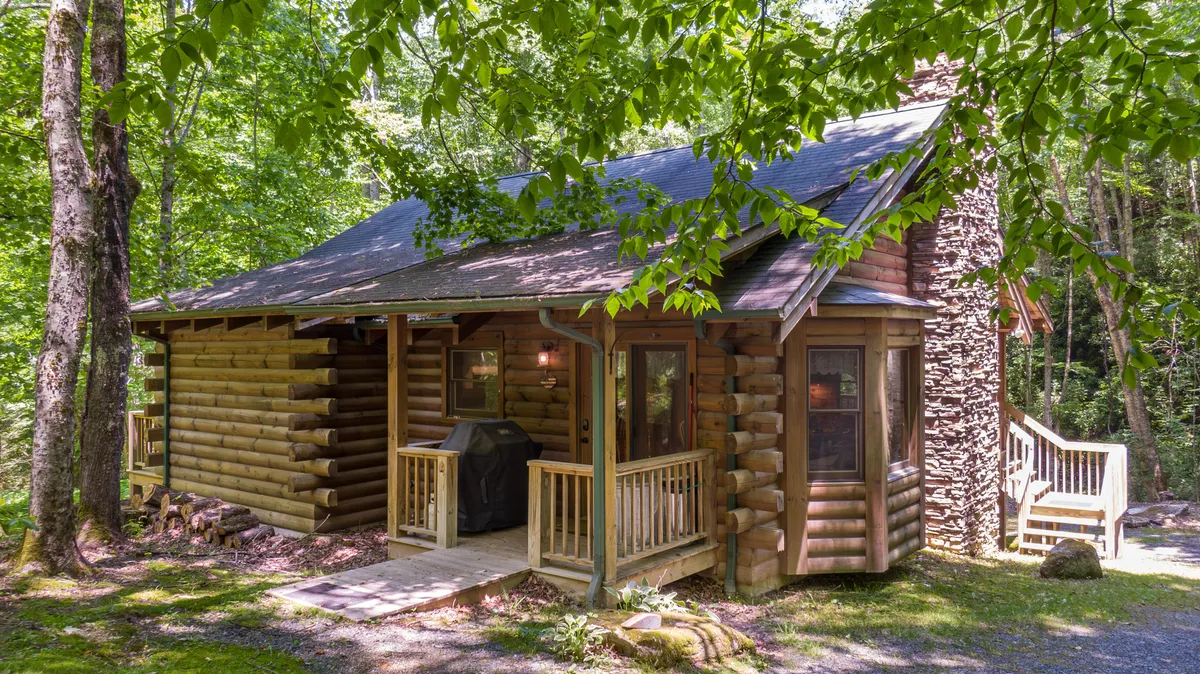 The image shows the log cabin’s inviting front porch, complete with a stone chimney, ramp access, and surrounded by mature trees. The porch is set up for relaxation and outdoor dining, hinting at grilling possibilities and easy access.