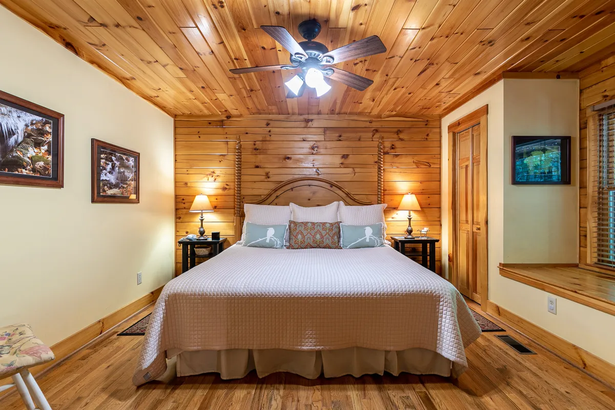 A serene, warmly lit bedroom featuring a king-size bed adorned with plush bedding, a wooden accent wall that adds rustic charm, side tables with softly glowing lamps, and decorative pillows. A window offers a view of the night sky, creating a peaceful atmosphere ideal for relaxation.