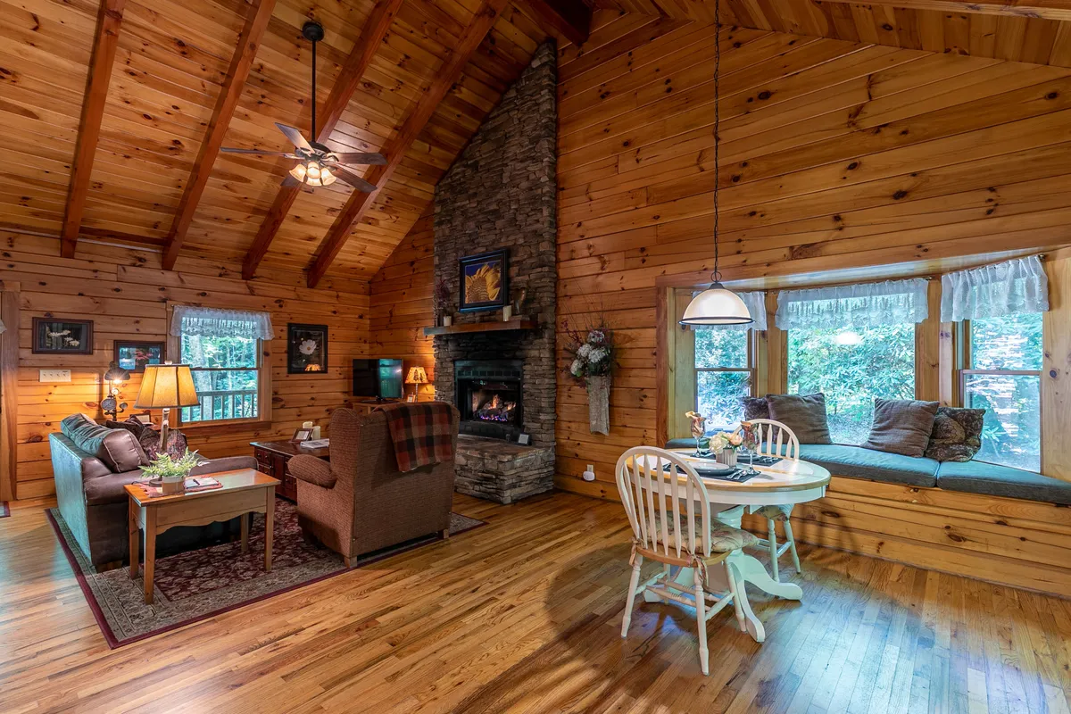 A spacious yet intimate living area showcasing a stone fireplace, exposed wooden beams, a cozy seating arrangement, and a charming window seat overlooking scenic wooded views. The arrangement invites guests to read, relax, or enjoy an intimate meal beside the fire or at the window.