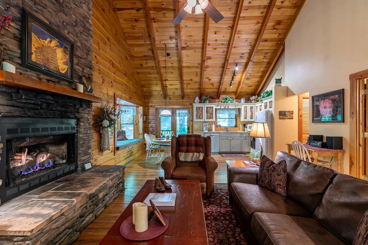 A broader view of the open-plan living area with a stone fireplace, exposed wooden beams, comfortable seating, and the kitchen area visible in the background. The scene is warm and welcoming, ideal for gatherings and shared meals.