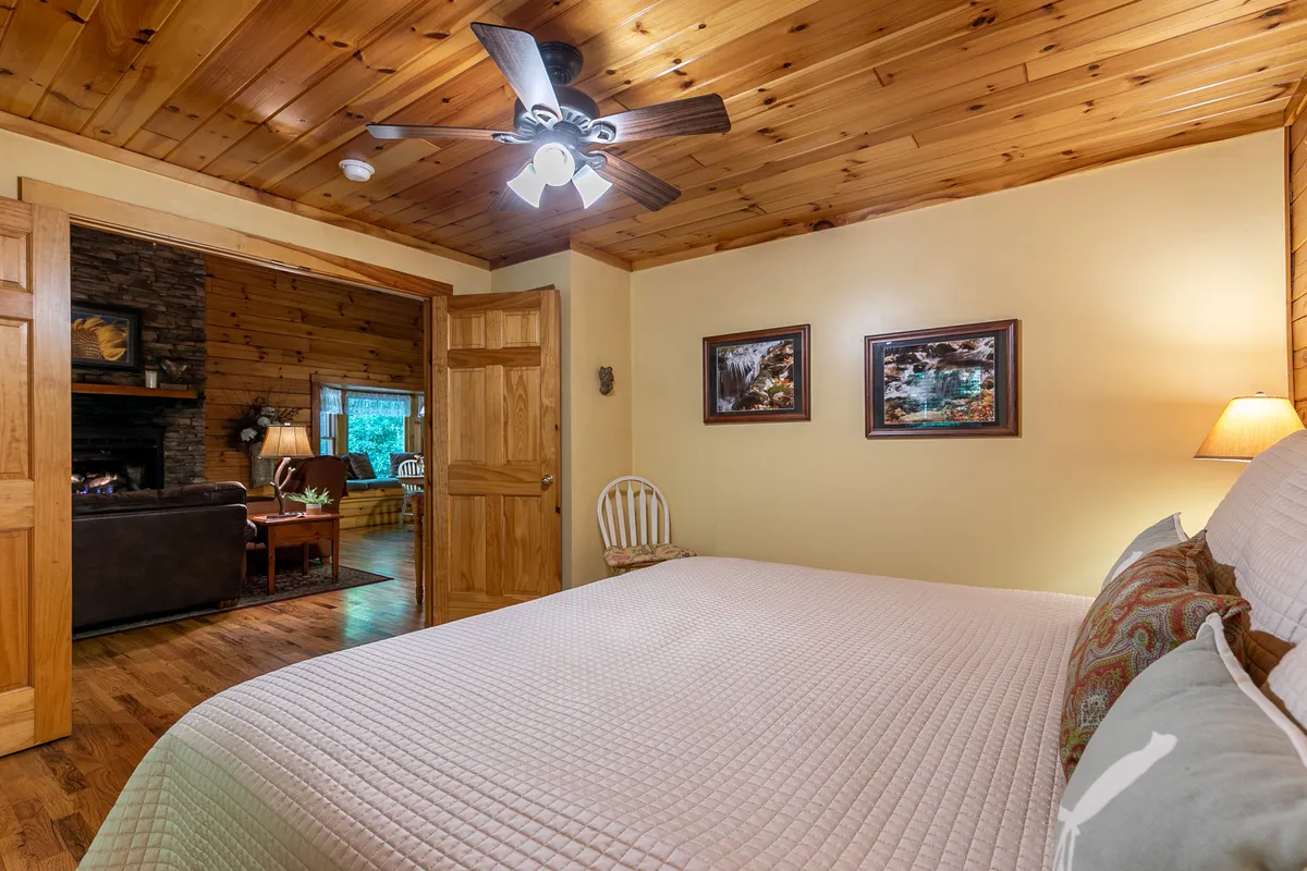 A welcoming bedroom with a warm wooden ceiling, a comfortable queen bed adorned with plush bedding, and tasteful artwork on the walls. The window offers a view into the living room, hinting at a cozy, communal space just beyond. The soft lighting and inviting textures create an atmosphere perfect for relaxation and unwinding after a day spent outdoors.