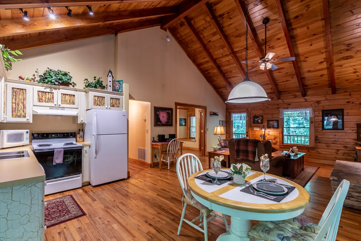 This image features a dining table set for two, rustic ceiling beams, a comfortable seating area, and glimpses of kitchen appliances in the background. The intimate table setting is accentuated by natural light and the cozy atmosphere of the adjoining living space.