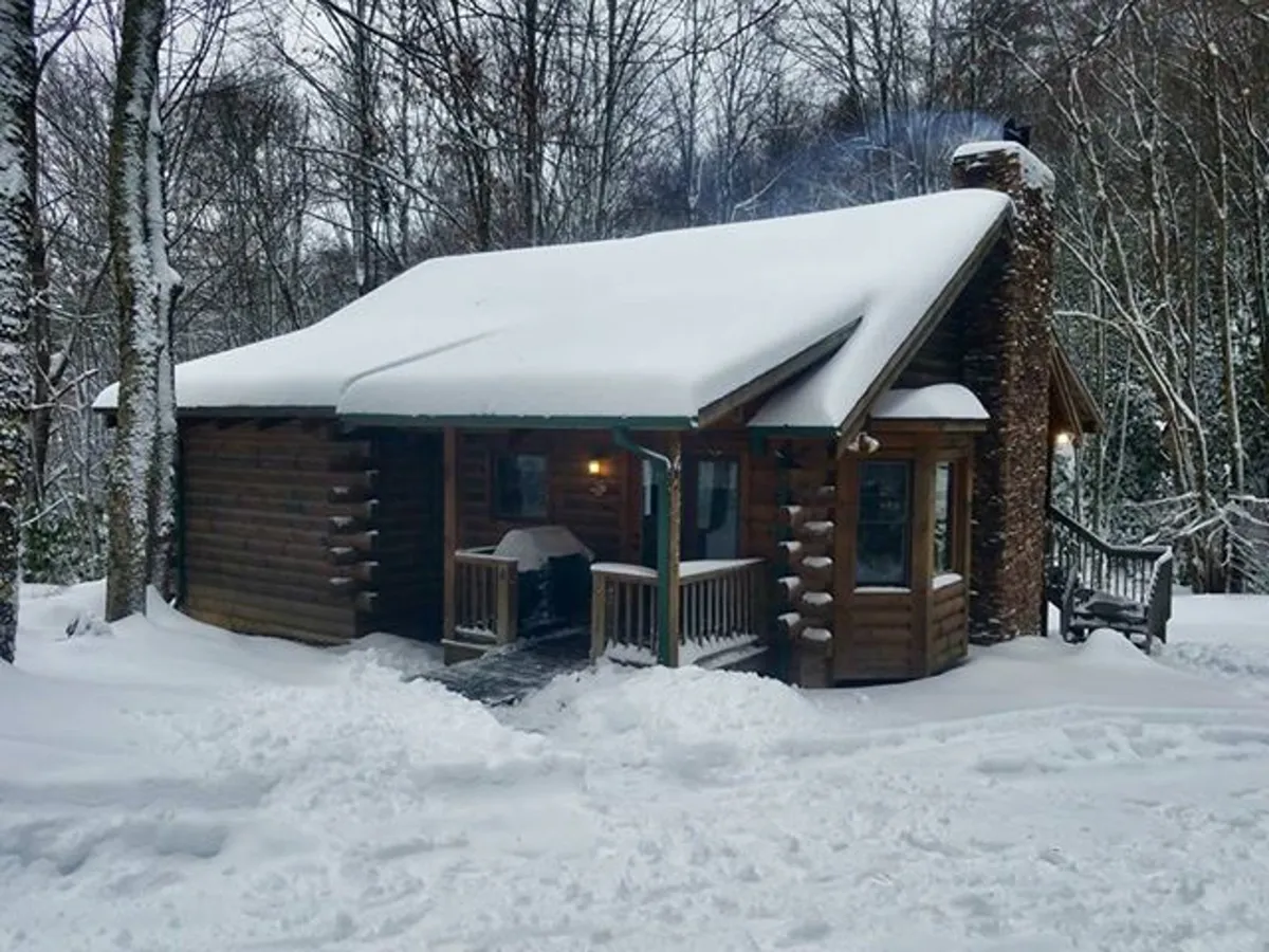 This image showcases the cabin's inviting exterior, featuring a rustic stone chimney and a snow-covered setting. The porch is visible, hinting at a cozy space to enjoy the outdoors even during winter.