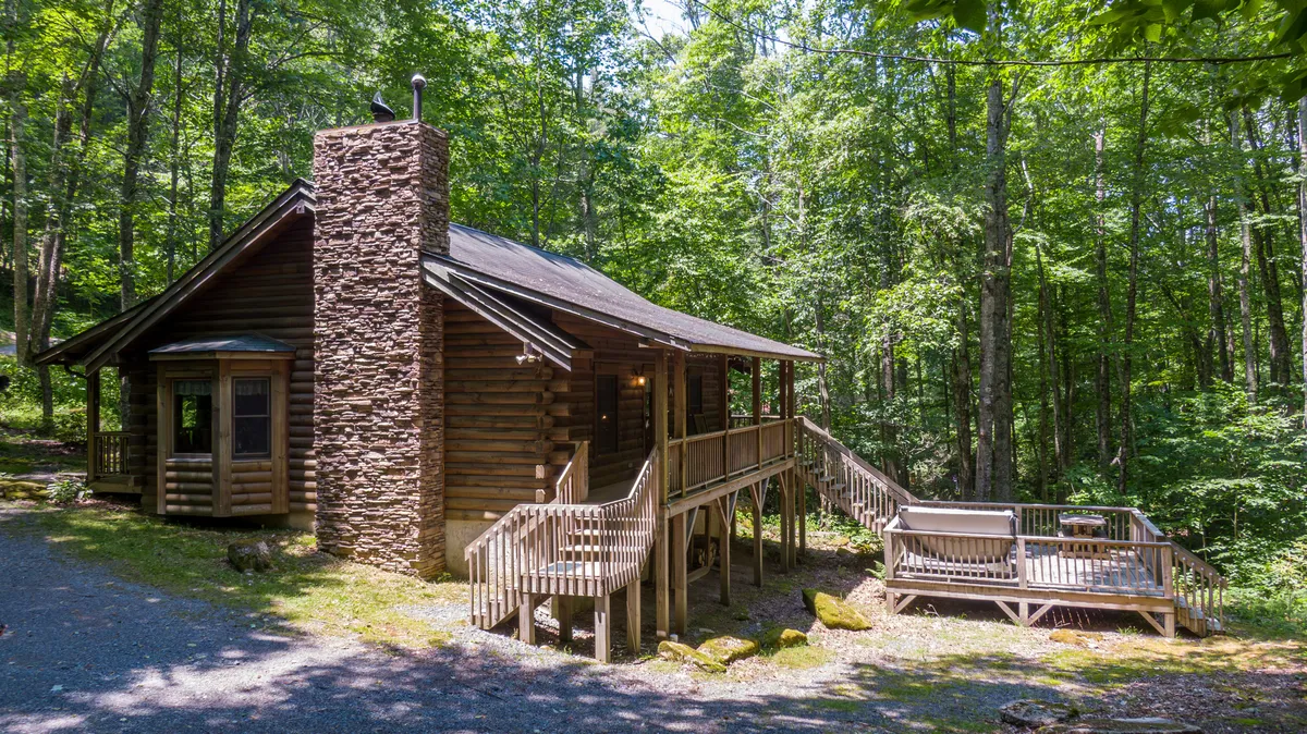 The image showcases a charming log cabin nestled amid towering trees, featuring a classic stone chimney, a welcoming wooden deck, and lush natural surroundings. Sunlight filters through the branches, casting a warm glow over the cabin, inviting guests to relax outdoors or explore the scenic beauty just steps away.