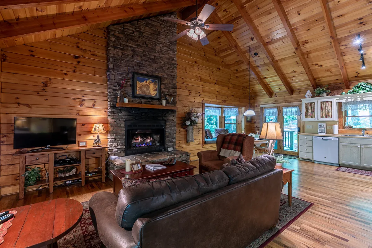 A warmly lit living room featuring a striking stone fireplace, a plush leather sofa, large windows that bathe the space in sunlight, and a rustic wooden ceiling. The scene conveys a blend of comfort and rustic style, with ample space to relax by the fire or gather with loved ones.